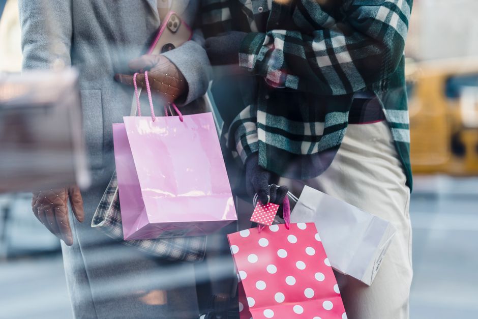 Crop faceless women in trendy outerwear carrying paper bags in hands and standing near shop window