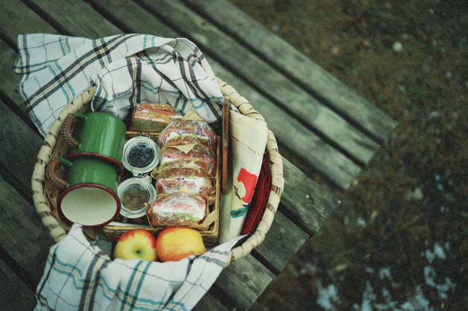 A classic wicker picnic basket open on a blanket outdoors, filled with food, glasses and cutlery