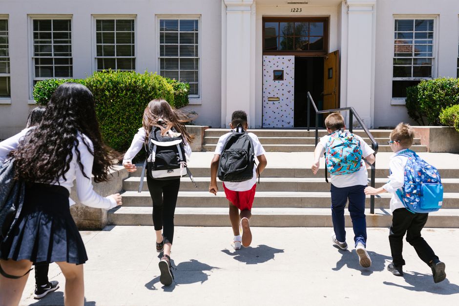Group of students with backpacks running into school, captured from behind, showing excitement.