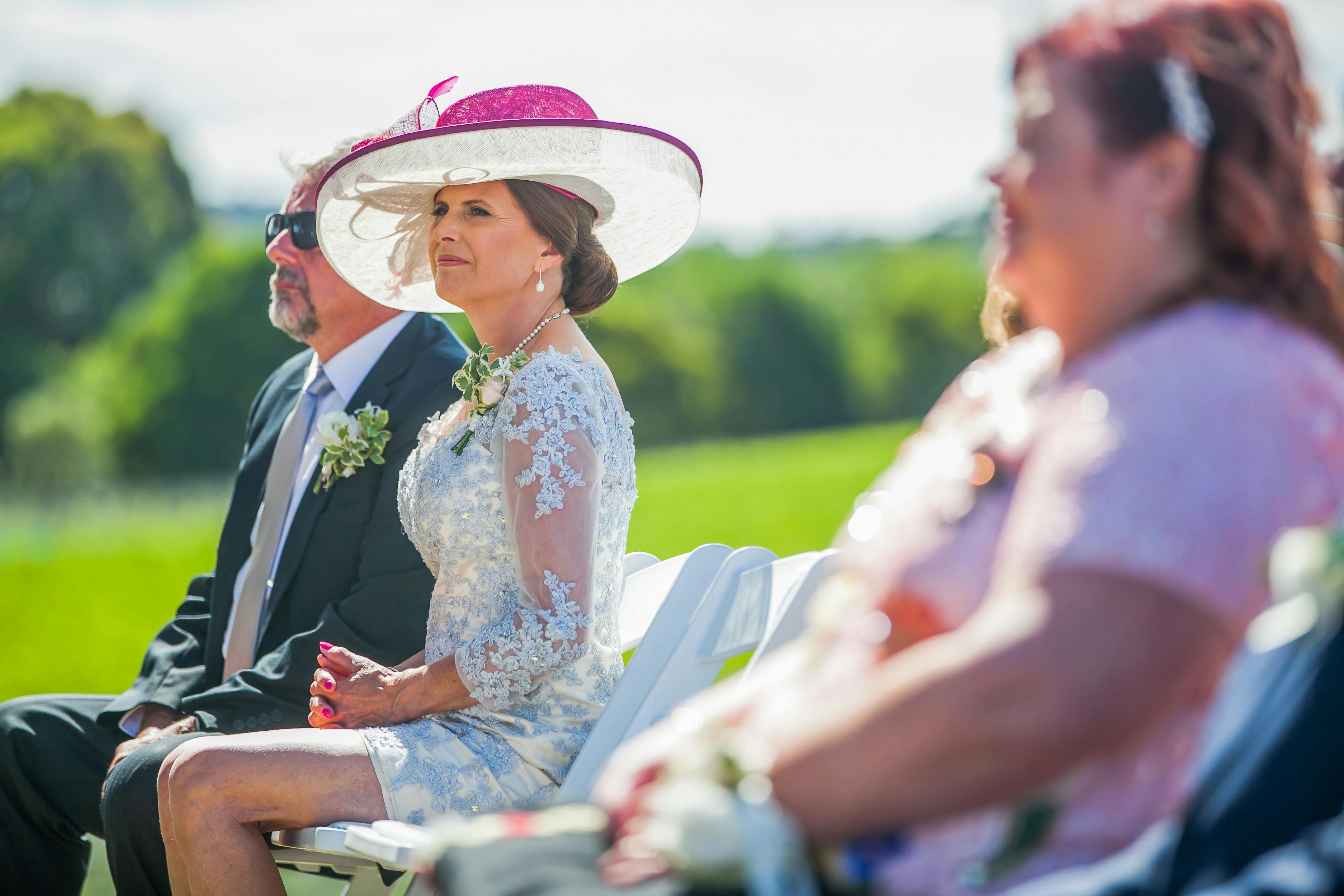 Side view of gorgeous middle aged female in elegant lace dress and hat sitting on chair near husband during outdoor wedding ceremony