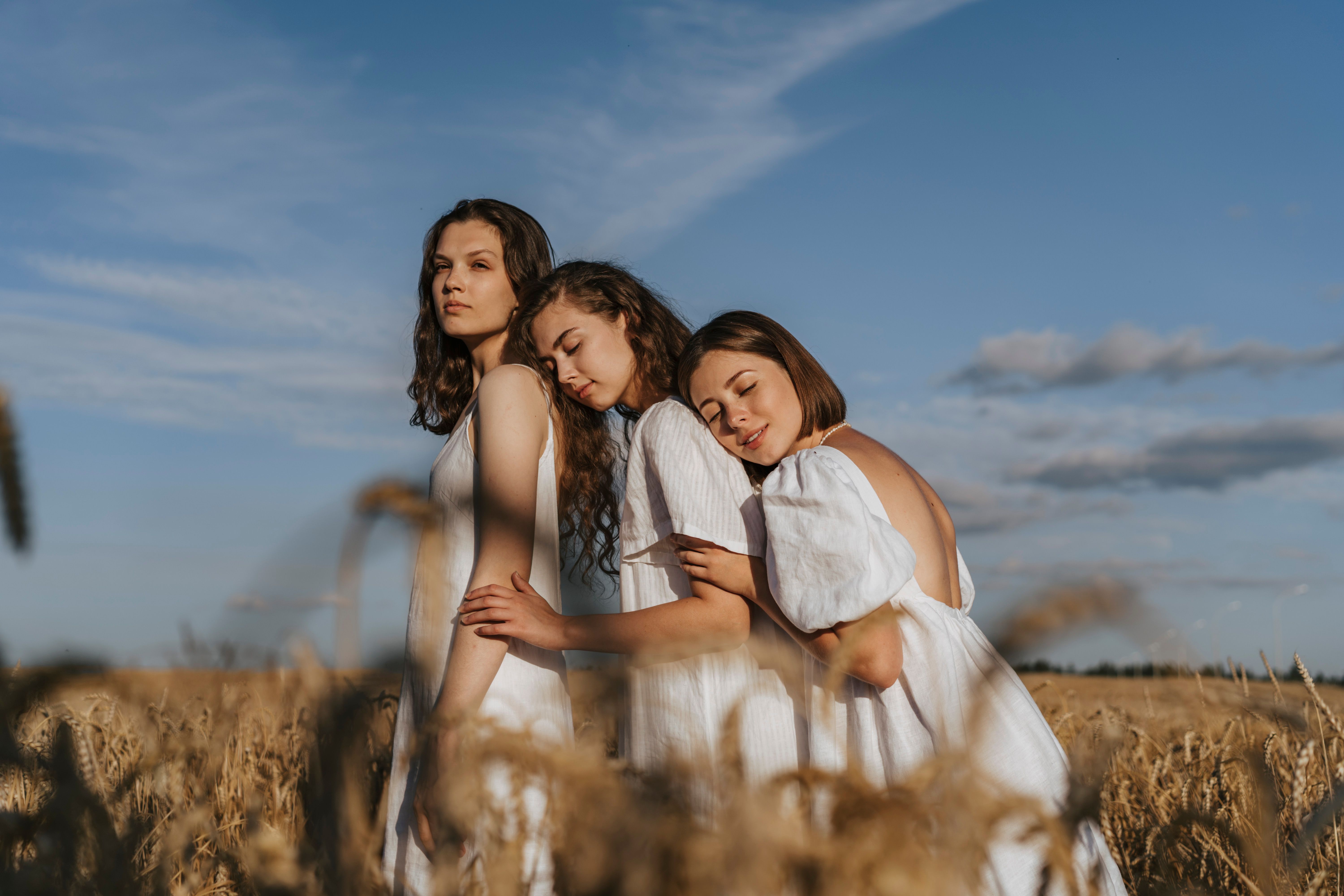 Three women in white dresses embracing in a sunlit wheat field capturing a serene moment of togetherness.
