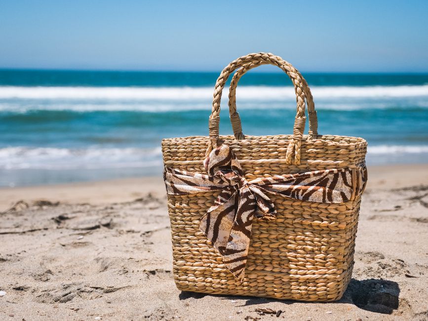 A woven straw tote bag on a sunny beach beside sunglasses and a sun hat