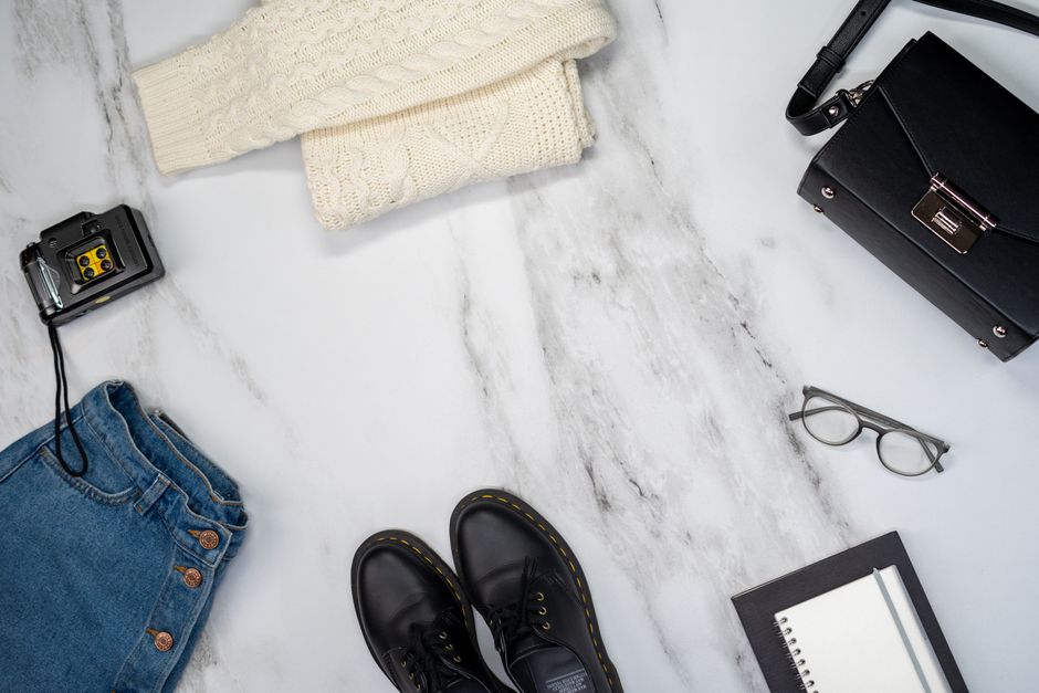 A flatlay of summer wardrobe essentials including sandals, sunglasses, a linen shirt and a straw bag arranged on a light background