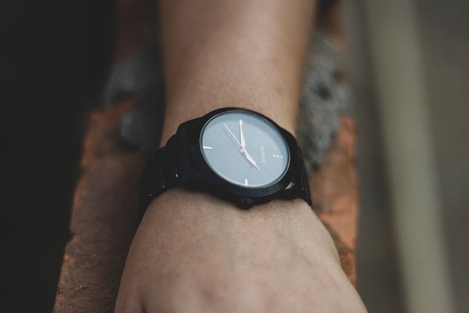 A person wearing a minimalist Scandinavian-style watch with a clean white dial and mesh strap against a summer outdoor background