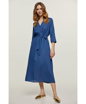 Dieses blaue Midikleid ist aus einem gewebten Leinenstoff gefertigt. Es hat einen Kragen und einen V-Ausschnitt mit einer Falte darunter. Es hat ¾ lange Ärmel mit Manschetten, die mit einem Kunststoffknopf Ton in Ton geschlossen werden. Es gibt einen Gürtel aus demselben Stoff und seitliche Innentaschen. Das Kleid ist in einer lockeren Silhouette gestylt.