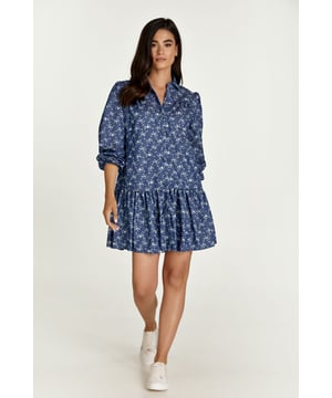 Este vestido floral índigo de manga larga está confeccionado en algodón tejido estilo denim. Tiene cuello y tapeta en la parte delantera que se abrocha con botones decorativos bicolor. Las mangas tienen puños de 3 cm que se abrochan con botón. Hay fruncidos en los hombros y 12 cm por debajo hay una costura con fruncidos debajo. A 30cm del dobladillo hay una costura con frunces.