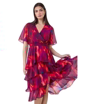 Abstract Print Chiffon Tiered Midi Dress from Roman. Discover pure elegance with this abstract print tiered midi dress from the Ariella collection. This wedding guest staple features a soft chiffon fabric with a flattering tiered silhouette and a v-neckline. Designed with a wrap front and shirred stretch waist for a fit and flare shape, this will be flattering for any formal event. The vibrant abstract print, split detail skirt, and short floaty sleeves add a touch of femininity to the finished look. Whether you're ready for a day at the races or you're Mother of the Bride, this dress is easy to pull on overhead and style with heels for timeless occasionwear.