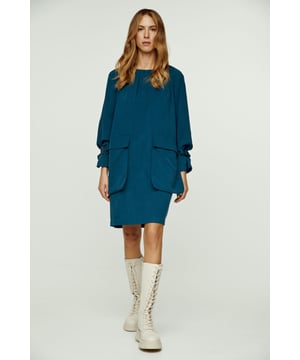 Este vestido azul petróleo está confeccionado en tencel tejido. Tiene mangas largas raglán y un amplio escote redondo con frunces. Una correa y una hebilla de plástico permiten fruncir las mangas en la muñeca. Hay dos bolsillos exteriores grandes y sueltos en la parte delantera de la prenda. El vestido se abrocha en la parte posterior con una cremallera oculta.