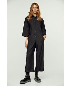 Dieser dunkelgraue Jumpsuit ist aus gewebtem Stretch-Denim-Stil gefertigt. Er hat Â¾ Ärmel und einen Kragen. Am Halsausschnitt befindet sich eine Knopfleiste mit schwarzen Kunststoffknöpfen. An den Seiten befinden sich Taschen mit je einer Gürtelschlaufe. Es hat einen 3 cm breiten Gürtel aus demselben Stoff, der durch zwei Gürtelschlaufen in der Taille geführt wird. Im Rücken befindet sich eine umgekehrte Falte. Der Jumpsuit hat eine Beinlänge von 7/8.