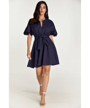 Este vestido azul oscuro está confeccionado en tejido de algodón bordado tono sobre tono. Tiene mangas cortas con doble volante y hombros caídos.  Tiene escote redondo con abertura en V en la parte delantera. Bajo el busto hay una costura curvada por debajo de la cual el vestido se abre en forma de cloche. En el interior, una cremallera oculta facilita la colocación del vestido. Está forrado. El vestido puede llevarse con o sin el cinturón del mismo tejido. El cinturón está incluido.