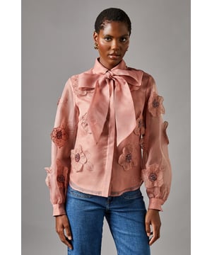 Exquisite organza blouse adorned with striking 3D flower appliqus
        Elegant collared neckline with feminine pussy bow detail
        Voluminous balloon sleeves for dramatic silhouette
        Semi-sheer finish with lined body for sophisticated coverage
        Button-through front design for classic styling
    
    This enchanting Coast blouse captures attention with its dimensional floral detailing and diaphanous organza fabric. The romantic balloon sleeves and graceful pussy bow create a silhouette that commands attention at any formal gathering. Style with wide-leg denim as shown for a contemporary twist on occasion dressing, or pair with a tailored pencil skirt and court shoes for wedding guest perfection. The translucent quality of the fabric creates an ethereal effect whilst the lined body ensures appropriate coverage. This versatile piece transitions beautifully from daytime events like the races to evening celebrations, making it an essential addition to your occasion wardrobe. The delicate craftsmanship and attention to detail reflect the sophisticated aesthetic Coast is renowned for.