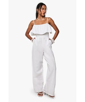 Feminine ruffle overlay adds a playful touch to this strappy jumpsuit
        Wide-leg silhouette creates an effortlessly chic and comfortable fit
        Practical side pockets for convenient storage of small essentials
        Cotton-linen blend fabric offers breathable comfort for all-day wear
        Adjustable thin straps allow for a customised fit to suit your preference
    
    This breezy cotton mix jumpsuit combines casual comfort with undeniable style. The delicate ruffle overlay creates movement and dimension, whilst the wide-leg design ensures a relaxed yet flattering silhouette. Perfect for summer gatherings, pair with flat sandals and a woven bag for daytime events as shown on our model. For evening casual occasions, add statement jewellery and metallic accessories to transform your look. The strappy neckline showcases your shoulders beautifully, making this jumpsuit ideal for warm-weather socialising. Versatile enough for family barbecues or casual brunches with friends, this jumpsuit offers the convenience of an all-in-one outfit with the comfort of natural fibres. The neutral tone makes it an excellent canvas for both minimalist and bold accessorising depending on your mood.