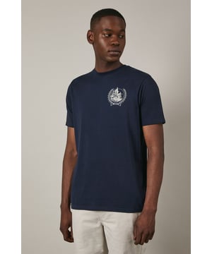 Soft jersey fabric for all-day comfort and breathability
        Classic crew neckline with ribbed trim for a neat finish
        Distinctive coastal ship emblem on chest for nautical appeal
        Short sleeves with clean-cut edges for a streamlined look
        Relaxed fit through the body offering ease of movement
    
    This Maine coastal print t-shirt brings maritime charm to your casual wardrobe. The understated design features a distinctive sailing ship emblem encircled by a laurel wreath, creating a subtle focal point against the navy background. Style it as shown with light-coloured shorts and white trainers for a fresh summer look perfect for beachside strolls or weekend brunches. For cooler evenings, layer under an unstructured blazer or light cotton overshirt. The versatile design transitions effortlessly from daytime casual to relaxed evening gatherings. The cotton jersey construction ensures this t-shirt remains comfortable throughout the day, making it an ideal choice for family outings, casual meet-ups with friends, or simply lounging at home with understated style.