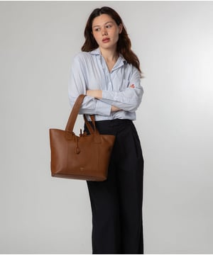 Handcrafted from natural leather with a soft to the touch pebble grain finish, the 'Carolina' tote bag from NiNa Amora is a must-have accessory. A unique tie top secures the central lined compartment which includes a zipped compartment which divides the design; perfect for maintaining an organised bag. Additional storage space is provided by a slip and zipped pocket on the interior, ideal for your phone, keys and any other essentials while out and about. Adorned with dual leather handles with a drop length of 25cm for a comfortable and versatile carrying options. Silver metal fittings and zips give this design a elegant look which is complemented the NiNa Amora X logo is silver foil embossed on the front and hanging charm for a luxurious finishing touch.
