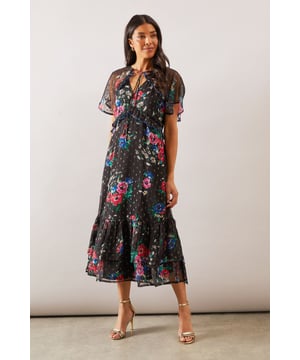 Stunning cape detail sleeves create a dramatic silhouette with elegant movement
        Flattering V-neckline with delicate tie detail for adjustable styling
        Sheer metallic overlay with vibrant floral print adds a touch of glamour
        Feminine midi length with tiered hem for added volume and sophistication
        Semi-sheer fabric creates a light, floaty effect perfect for special occasions
    
    This enchanting Wallis midi dress combines sophistication with contemporary flair. The striking cape sleeves create a graceful drape whilst the metallic printed overlay catches the light beautifully as you move. The flattering V-neckline frames the dcolletage perfectly, whilst the tiered hem adds a touch of drama to your silhouette. Style with strappy metallic heels as shown for formal events such as weddings or race days. For evening soires, add statement jewellery and a clutch bag to complete your ensemble. The versatile design transitions effortlessly from daytime celebrations to evening gatherings, making it a wardrobe essential for the woman who appreciates timeless elegance with a modern twist. This dress promises to make you feel both comfortable and confidently glamorous for any special occasion.
