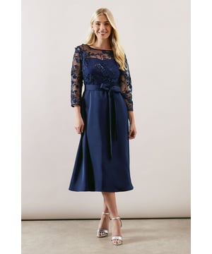 Elegant mesh overlay bodice with intricate floral embroidery detail
        Sophisticated crew neckline with three-quarter length sheer sleeves
        Flattering fit and flare silhouette that cinches at the waist
        Luxurious satin skirt that falls to a graceful midi length
        Feminine self-tie belt to accentuate the waistline
    
    This enchanting occasion dress combines the delicate beauty of embroidered mesh with the refined elegance of a satin skirt. The bodice features exquisite floral detailing that creates a striking contrast against the smooth, flowing fabric below. Perfect for formal gatherings, this Wallis dress offers both sophistication and comfort for the discerning woman. Style with metallic strappy heels as shown for a polished wedding guest ensemble, or pair with a structured clutch and delicate jewellery for race day glamour. The versatile midi length works beautifully for daytime ceremonies or evening celebrations, while the fit and flare shape flatters the figure with timeless appeal. This dress strikes the perfect balance between contemporary design and classic occasion wear, making it a standout choice for your most memorable events.