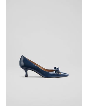 The Alma eyelet bow courts, blue patent pumps crafted from 100% leather, feature a pointed toe, kitten heel, and eyelet bow detail with metal ends. Versatile and stylish, they complement tailored trousers and elegant dresses, suitable for any smart-casual occasion. Made in Spain, ideal for brunches, office parties, or an evening out with friends.