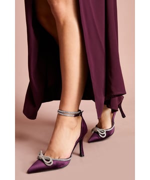 Elegant satin court shoes featuring a striking diamante bow detail at the toe
        Slender ankle strap with adjustable buckle fastening for a secure fit
        Pointed toe silhouette for a sophisticated, elongating effect
        High stiletto heel measuring between 7-9cm for a glamorous lift
        Versatile design perfect for formal occasions and evening events
    
    The Krissy Satin Diamante Bow Court Shoes manifest timeless elegance with a contemporary twist. The luxurious satin finish creates a refined canvas for the show-stopping diamante bow that adorns the pointed toe, catching the light with every step. Designed with a high stiletto heel that measures between 7-9cm, these shoes offer the perfect balance of height and wearability for special occasions. Style them with a tailored midi dress for weddings or pair with a sleek jumpsuit for race day sophistication. The delicate ankle strap not only provides additional support but also adds a feminine touch to the silhouette. These Coast heels are the ultimate finishing touch for any formal ensemble, whether you're attending a graduation ceremony, black-tie event, or celebrating a milestone occasion. Their refined aesthetic ensures they'll remain a treasured addition to your occasion wardrobe for years to come.