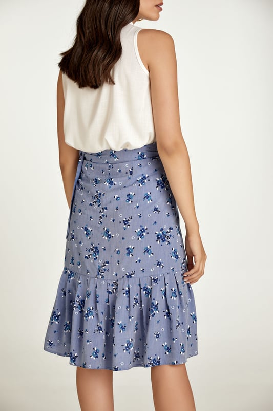 Esta falda cruzada floral está confeccionada en tejido de viscosa estilo denim. La falda se abrocha con una corbata de 4,5 cm del mismo tejido que pasa a través de un agujero en la costura lateral, justo debajo de la cintura. La falda tiene un ribete de volantes de 26 cm por todas partes. Tiene un estilo sutil con una silueta de línea A y llega hasta la rodilla.