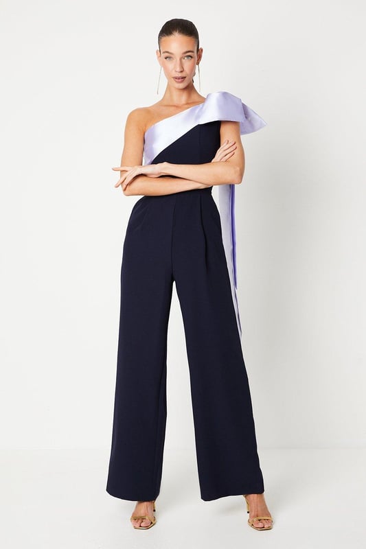 One-shoulder design with a dramatic bow detail
Wide-leg jumpsuit silhouette with a flattering waist tie
crepe fabric with a luxurious drape
Sleeveless bodice with a fitted waist
Full-length wide legs for an elegant, flowing look

Exuding sophistication and glamour, this jumpsuit is a showstopping choice for formal occasions. The one-shoulder neckline with a striking bow detail adds a touch of drama, while the crepe fabric drapes beautifully for a refined look. Style it with statement jewellery and heels for a wedding or evening event, or pair it with a fascinator and clutch for a day at the races. With its flattering waist tie and flowing wide legs, this jumpsuit is sure to turn heads and make a lasting impression.