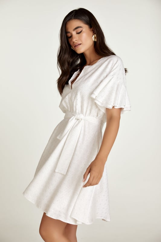 Este vestido blanco está confeccionado en tejido de viscosa bordado tono sobre tono. Tiene mangas cortas con doble volante y hombros caídos. Tiene escote redondo con abertura en V en la parte delantera. Debajo del busto hay una costura curva bajo la cual el vestido se abre en forma de campana. Hay una cremallera oculta en el interior que permite ponerse el vestido fácilmente. Está forrado. El vestido se puede llevar con o sin cinturón que está confeccionado en el mismo tejido. El cinturón está incluido.