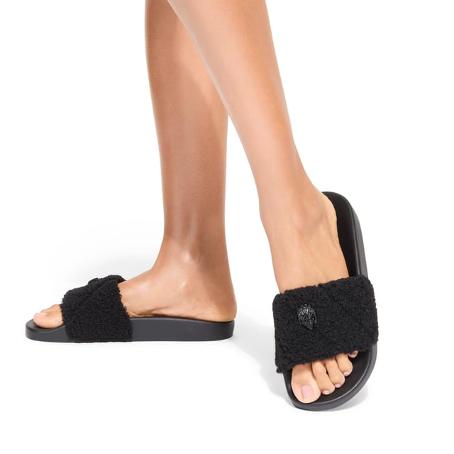 The Kurt Geiger Meena Teddy is a slide on sandal with a black faux shearling strap. There is a black eagle head sat on the strap that is embellished with crystals.