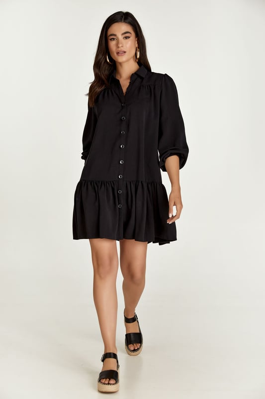 Este vestido negro de manga larga está confeccionado en tencel tejido. Tiene cuello y tapeta en la parte delantera que se abrocha con botones decorativos de plástico negro. Las mangas tienen puños de 3 cm que se abrochan con botón. Hay fruncidos en los hombros y 12 cm por debajo hay una costura con fruncidos debajo. A 30cm del dobladillo hay una costura con frunces.
