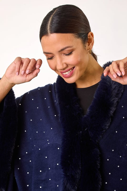 Hotfix Faux Fur Trim Cape from Roman. This style is ONE size.  Wrap up in style this Autumn and Winter with this all over hotfix faux fur trim cape. In a classic black hue with dazzling silver hotfix detailing, this cosy cape is the perfect layering piece. Simply throw on over a t-shirt and jeans for an effortlessly chic and stylish look.
