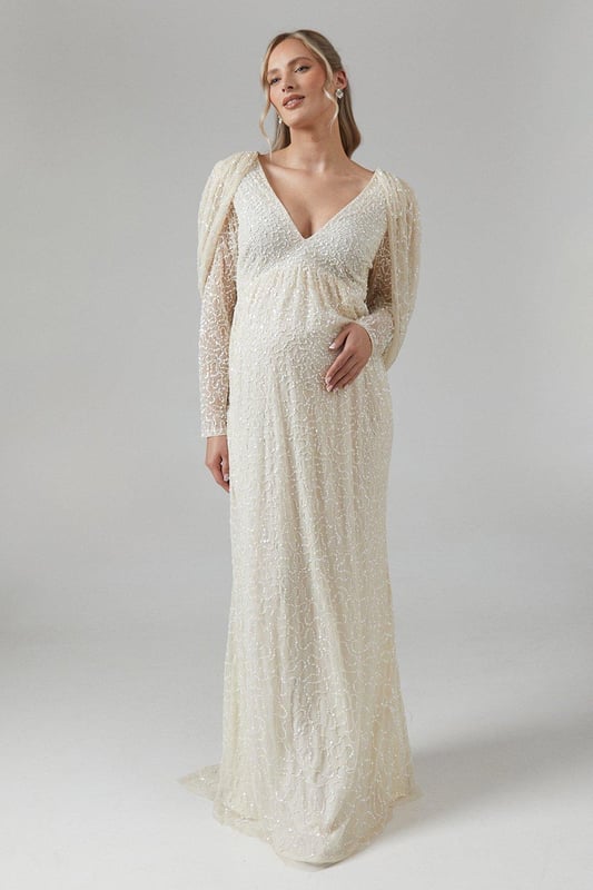 Exquisite hand-stitched embellishments for a luxurious finish
        Elegant V-neckline complemented by long sleeves
        Stunning cowl back detail for added sophistication
        Floor-length design with a graceful silhouette
        Maternity-friendly cut to accommodate and flatter your changing shape
    
    Radiate bridal elegance in this captivating maternity wedding dress from Coast. The hand-stitched embellishments create a dazzling effect, perfect for catching the light as you walk down the aisle. The V-neckline and long sleeves offer a timeless appeal, while the cowl back adds a touch of drama to your ensemble. Designed with the expectant bride in mind, this gown gracefully accommodates your changing silhouette, ensuring comfort throughout your special day. Style with delicate drop earrings and a simple bracelet to let the dress take centre stage. For a complete bridal look, consider a floor-length veil and ivory satin heels. This gown is not just for the ceremony; it transitions beautifully to the reception, allowing you to dance the night away in style and comfort.