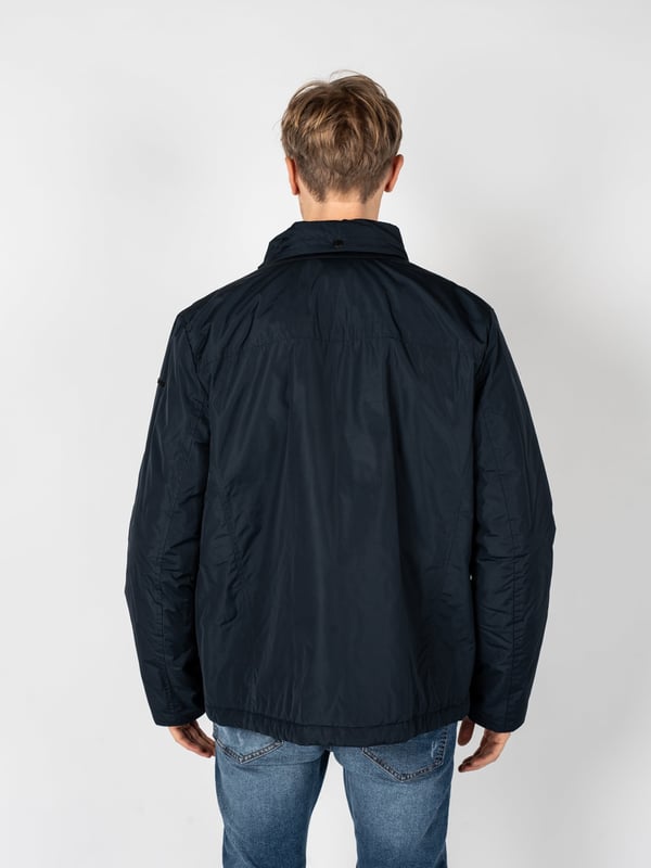 Un diseño sencillo es la solución perfecta para aquellos que valoran la clásica y el minimalismo, al mismo tiempo que desean verse estilosos y elegantes en cualquier situación.

El cuello alto añade un carácter moderno al conjunto, a la vez que ofrece protección adicional contra el viento y el frío, lo que hace que esta prenda sea una excelente elección para las condiciones meteorológicas cambiantes.

Con una capucha oculta en el cuello, puedes estar preparado para las lluvias inesperadas, y su ingenioso diseño permite que se guarde fácilmente cuando no es necesaria, manteniendo un aspecto elegante.

Se abrocha con una cremallera logotipada y botones a presión, lo que no solo añade un estilo único, sino que también proporciona protección adicional contra el frío, destacando al mismo tiempo la atención a los detalles.

En el frente, cuatro bolsillos, dos con cremallera y dos con botones a presión, ofrecen mucho espacio para guardar los artículos esenciales y su variedad permite una mejor organización.

En la manga izquierda, un detalle de la marca constituye un sutil, pero claro refuerzo del prestigio de la marca, agregando un carácter exclusivo a la prenda y distinguiéndola de las demás.

Fabricada con tela de alta calidad, transpirable y resistente al agua, lo que garantiza no solo comodidad al usarla, sino también protección contra las condiciones atmosféricas adversas, haciendo que sea una elección ideal para cualquier época del año.