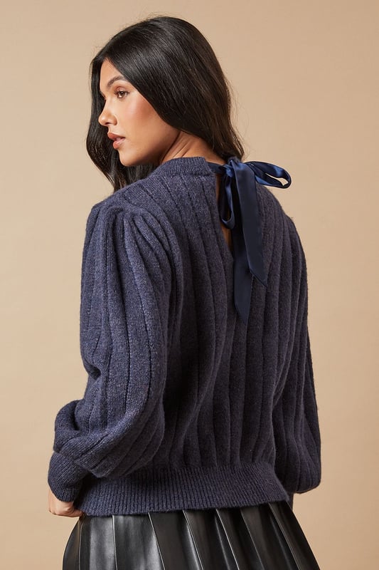 Ribbed knit texture for added visual interest
        Elegant bow detail at the back for a feminine touch
        Voluminous balloon sleeves create a striking silhouette
        Crew neckline for a classic finish
        Versatile design suitable for various casual occasions
    
    This Oasis Bow Back Ribbed Jumper effortlessly combines comfort with style, making it a must-have addition to your wardrobe. The ribbed knit texture adds depth and interest to the design, while the charming bow detail at the back introduces a delightful feminine flair. The jumper's standout feature is undoubtedly its voluminous balloon sleeves, which create a dramatic and eye-catching silhouette. For a casual yet chic ensemble, pair this jumper with a pleated leather-look skirt and knee-high boots, as shown in the image. This versatile piece is perfect for family gatherings, brunch dates, or casual outings with friends. To dress it up for evening drinks, consider swapping the boots for heeled ankle booties and adding statement earrings. The jumper's adaptable nature ensures it will become a go-to item in your wardrobe, easily transitioning from day to night with simple styling tweaks.