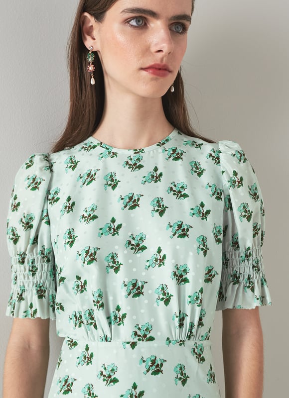 The Tabitha dress is crafted from a silk-blend spot jacquard in morning mist green with a 1930's hand painted primula print reimagined from vintage archives. It has a round neck, puff shoulders, short shirred sleeves, pleating at the bust, a floaty shape and a dipped midi-length hem.