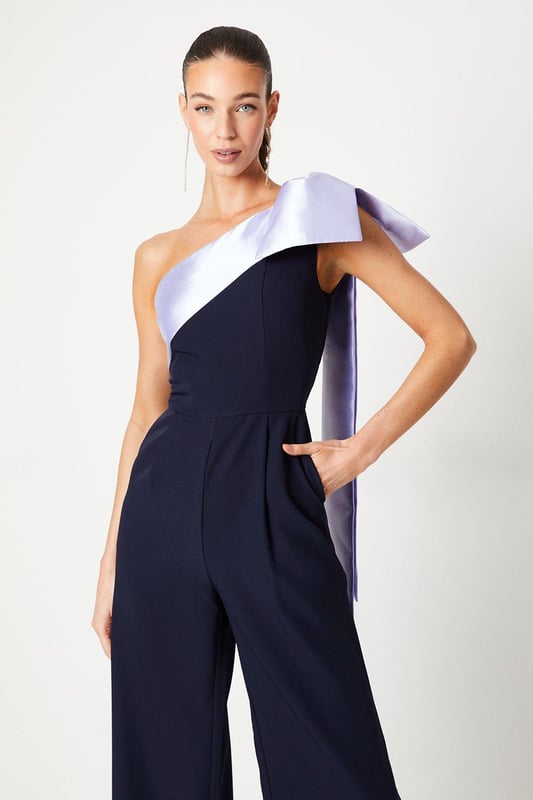 One-shoulder design with a dramatic bow detail
Wide-leg jumpsuit silhouette with a flattering waist tie
crepe fabric with a luxurious drape
Sleeveless bodice with a fitted waist
Full-length wide legs for an elegant, flowing look

Exuding sophistication and glamour, this jumpsuit is a showstopping choice for formal occasions. The one-shoulder neckline with a striking bow detail adds a touch of drama, while the crepe fabric drapes beautifully for a refined look. Style it with statement jewellery and heels for a wedding or evening event, or pair it with a fascinator and clutch for a day at the races. With its flattering waist tie and flowing wide legs, this jumpsuit is sure to turn heads and make a lasting impression.