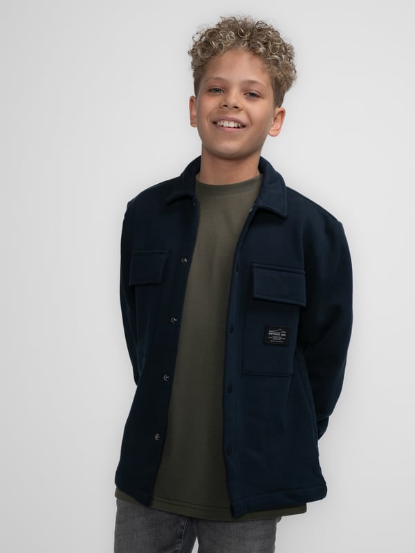 Overshirt aus Sweatstoff mit langen Ärmeln. Dieses Hemd verfügt über einen klassischen Kragen, einen Druckknopfverschluss, zwei Brusttaschen mit Druckknopfverschluss und ein Emblem auf einer Brusttasche. Das Overshirt ist mit Druckknöpfen an den Ärmeln versehen. Haben Sie Schwierigkeiten, sich zwischen zwei Größen zu entscheiden? Dann wählen Sie die größere Größe. Sweatstoff, klassischer Kragen, Druckknopfverschluss, zwei Brusttaschen, 100 % Baumwolle.