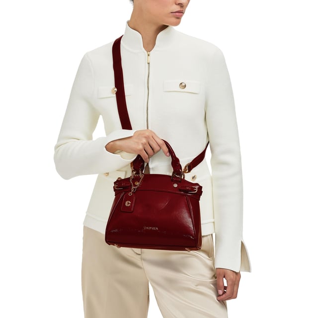 This Carvela Mini Valentina Tote bag features a wine red exterior in a trapeze style. There are two buckled straps across each side of the bag and branded gold tone hardware across the bag. Dimensions: 19cm (H), 22cm (L), 10cm (D). Strap length: 111cm.