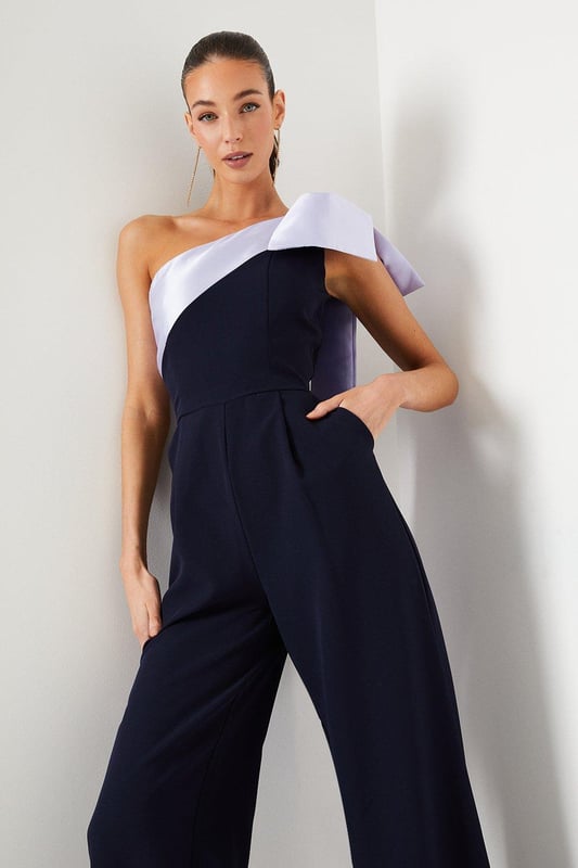 One-shoulder design with a dramatic bow detail
Wide-leg jumpsuit silhouette with a flattering waist tie
crepe fabric with a luxurious drape
Sleeveless bodice with a fitted waist
Full-length wide legs for an elegant, flowing look

Exuding sophistication and glamour, this jumpsuit is a showstopping choice for formal occasions. The one-shoulder neckline with a striking bow detail adds a touch of drama, while the crepe fabric drapes beautifully for a refined look. Style it with statement jewellery and heels for a wedding or evening event, or pair it with a fascinator and clutch for a day at the races. With its flattering waist tie and flowing wide legs, this jumpsuit is sure to turn heads and make a lasting impression.