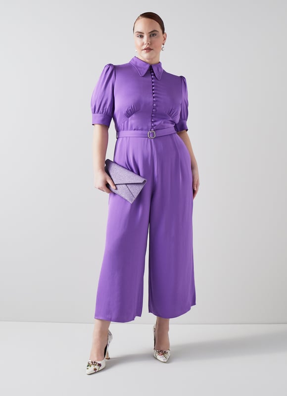 LK Bennett Aldous Dresses, Wisteria The Aldous jumpsuit in the Royal Ascot x LKB collection is crafted from a silky fabric in wisteria purple. It has a long rounded collar, puff shoulder short sleeves, a half placket of covered buttons, a diamante buckle belt, and wide leg cropped trousers.