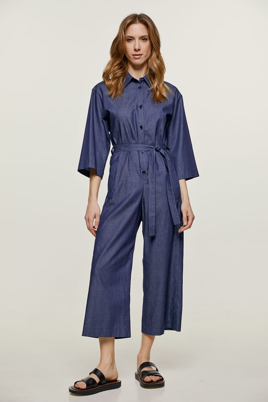 Deze indigo jumpsuit is gemaakt van katoen. Het heeft ¾ mouwen en een kraag. Er is een halssluiting met zwarte plastic knopen. Er zijn zakken aan de zijkanten met elk een riemlus. Het heeft een 3 cm brede ceintuur van dezelfde stof die door twee ceintuurlussen in de taille loopt. Aan de achterkant zit een omgekeerde plooi. De jumpsuit heeft een beenlengte van 7/8.