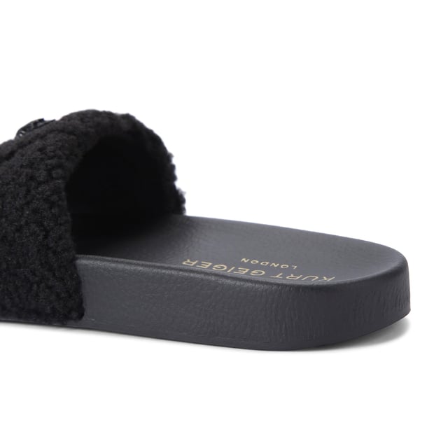 The Kurt Geiger Meena Teddy is a slide on sandal with a black faux shearling strap. There is a black eagle head sat on the strap that is embellished with crystals.