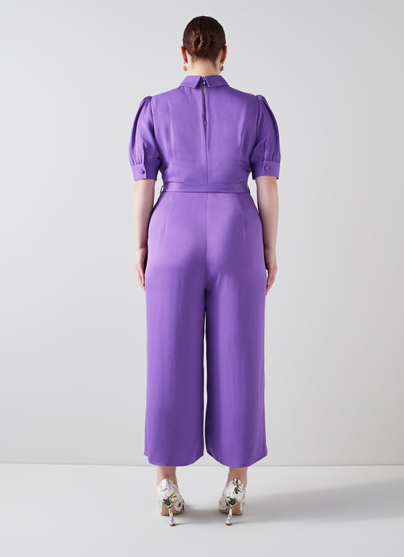 LK Bennett Aldous Dresses, Wisteria The Aldous jumpsuit in the Royal Ascot x LKB collection is crafted from a silky fabric in wisteria purple. It has a long rounded collar, puff shoulder short sleeves, a half placket of covered buttons, a diamante buckle belt, and wide leg cropped trousers.