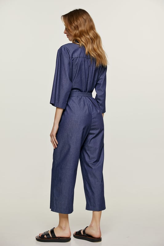 Deze indigo jumpsuit is gemaakt van katoen. Het heeft ¾ mouwen en een kraag. Er is een halssluiting met zwarte plastic knopen. Er zijn zakken aan de zijkanten met elk een riemlus. Het heeft een 3 cm brede ceintuur van dezelfde stof die door twee ceintuurlussen in de taille loopt. Aan de achterkant zit een omgekeerde plooi. De jumpsuit heeft een beenlengte van 7/8.