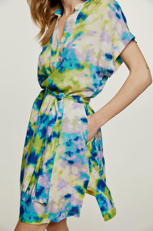 Deze zomerjurk met print is gemaakt van geweven viscose. Het is mouwloos en heeft een boord bij de armsgaten in dezelfde stof. Het heeft een mandarijnkraag van 2 cm en een sluiting van 4 cm bij de halslijn aan de voorkant om een V-vorm te creëren. Aan elke kant zit een binnenzak.  De jurk heeft grote splitten van 20 cm aan elke kant. De jurk kan worden gedragen met de riem of zonder als u een meer ontspannen zakjurkstijl wilt creëren. De riem in dezelfde stof is inbegrepen. Dit kledingstuk is gestileerd in een recht los silhouet.