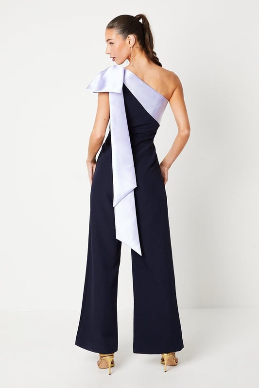 One-shoulder design with a dramatic bow detail
Wide-leg jumpsuit silhouette with a flattering waist tie
crepe fabric with a luxurious drape
Sleeveless bodice with a fitted waist
Full-length wide legs for an elegant, flowing look

Exuding sophistication and glamour, this jumpsuit is a showstopping choice for formal occasions. The one-shoulder neckline with a striking bow detail adds a touch of drama, while the crepe fabric drapes beautifully for a refined look. Style it with statement jewellery and heels for a wedding or evening event, or pair it with a fascinator and clutch for a day at the races. With its flattering waist tie and flowing wide legs, this jumpsuit is sure to turn heads and make a lasting impression.