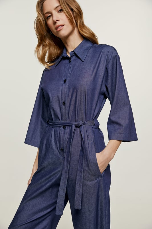Deze indigo jumpsuit is gemaakt van katoen. Het heeft ¾ mouwen en een kraag. Er is een halssluiting met zwarte plastic knopen. Er zijn zakken aan de zijkanten met elk een riemlus. Het heeft een 3 cm brede ceintuur van dezelfde stof die door twee ceintuurlussen in de taille loopt. Aan de achterkant zit een omgekeerde plooi. De jumpsuit heeft een beenlengte van 7/8.