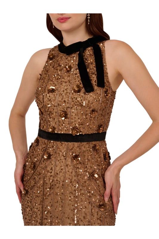 Make a bold and sparkling statement when you slip into this velvet and beaded gown. This formal dress features a halter bodice with velvet bow neckline, a velvet-trimmed waist, and a column skirt. Made with knit mesh and covered in decadent sequins and beading all over, this evening gown is trimmed with 3D sequin flowers. A hidden zipper closure at the centre back completes this column gown. A feminine look for a black-tie gala or formal attire wedding, this beaded dress will impress.