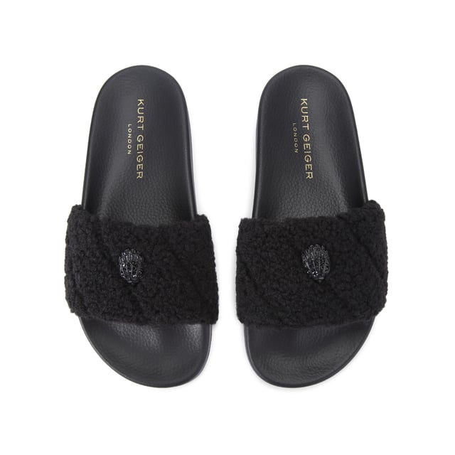 The Kurt Geiger Meena Teddy is a slide on sandal with a black faux shearling strap. There is a black eagle head sat on the strap that is embellished with crystals.