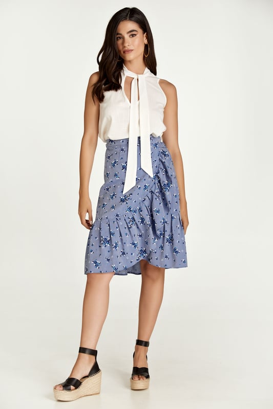 Esta falda cruzada floral está confeccionada en tejido de viscosa estilo denim. La falda se abrocha con una corbata de 4,5 cm del mismo tejido que pasa a través de un agujero en la costura lateral, justo debajo de la cintura. La falda tiene un ribete de volantes de 26 cm por todas partes. Tiene un estilo sutil con una silueta de línea A y llega hasta la rodilla.