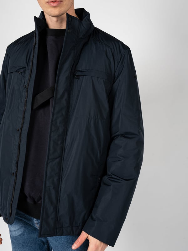 Un diseño sencillo es la solución perfecta para aquellos que valoran la clásica y el minimalismo, al mismo tiempo que desean verse estilosos y elegantes en cualquier situación.

El cuello alto añade un carácter moderno al conjunto, a la vez que ofrece protección adicional contra el viento y el frío, lo que hace que esta prenda sea una excelente elección para las condiciones meteorológicas cambiantes.

Con una capucha oculta en el cuello, puedes estar preparado para las lluvias inesperadas, y su ingenioso diseño permite que se guarde fácilmente cuando no es necesaria, manteniendo un aspecto elegante.

Se abrocha con una cremallera logotipada y botones a presión, lo que no solo añade un estilo único, sino que también proporciona protección adicional contra el frío, destacando al mismo tiempo la atención a los detalles.

En el frente, cuatro bolsillos, dos con cremallera y dos con botones a presión, ofrecen mucho espacio para guardar los artículos esenciales y su variedad permite una mejor organización.

En la manga izquierda, un detalle de la marca constituye un sutil, pero claro refuerzo del prestigio de la marca, agregando un carácter exclusivo a la prenda y distinguiéndola de las demás.

Fabricada con tela de alta calidad, transpirable y resistente al agua, lo que garantiza no solo comodidad al usarla, sino también protección contra las condiciones atmosféricas adversas, haciendo que sea una elección ideal para cualquier época del año.