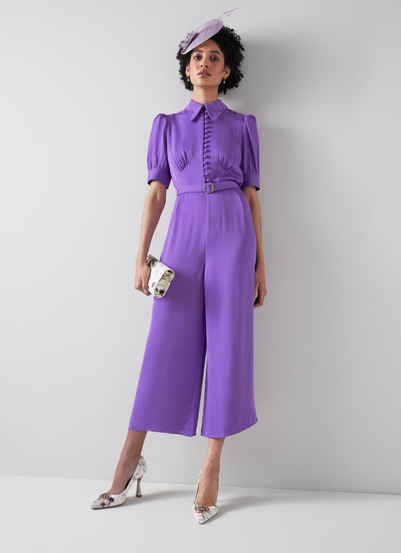 LK Bennett Aldous Dresses, Wisteria The Aldous jumpsuit in the Royal Ascot x LKB collection is crafted from a silky fabric in wisteria purple. It has a long rounded collar, puff shoulder short sleeves, a half placket of covered buttons, a diamante buckle belt, and wide leg cropped trousers.