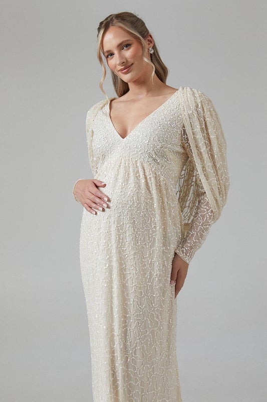 Exquisite hand-stitched embellishments for a luxurious finish
        Elegant V-neckline complemented by long sleeves
        Stunning cowl back detail for added sophistication
        Floor-length design with a graceful silhouette
        Maternity-friendly cut to accommodate and flatter your changing shape
    
    Radiate bridal elegance in this captivating maternity wedding dress from Coast. The hand-stitched embellishments create a dazzling effect, perfect for catching the light as you walk down the aisle. The V-neckline and long sleeves offer a timeless appeal, while the cowl back adds a touch of drama to your ensemble. Designed with the expectant bride in mind, this gown gracefully accommodates your changing silhouette, ensuring comfort throughout your special day. Style with delicate drop earrings and a simple bracelet to let the dress take centre stage. For a complete bridal look, consider a floor-length veil and ivory satin heels. This gown is not just for the ceremony; it transitions beautifully to the reception, allowing you to dance the night away in style and comfort.